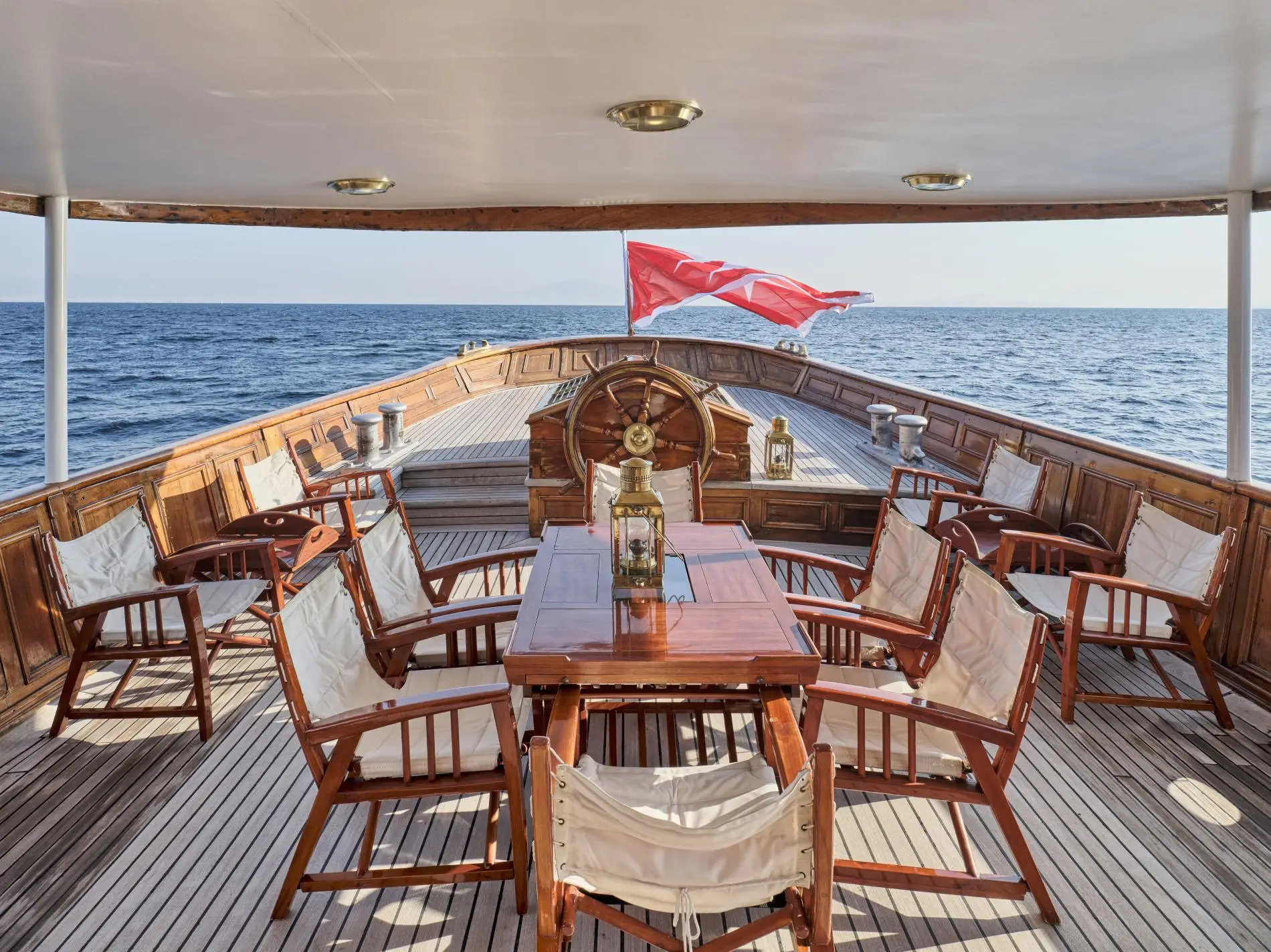 Madiz's aft deck under an awning, with folding chairs, a varnished table, the ship's wheel and the Maltese ensign astern