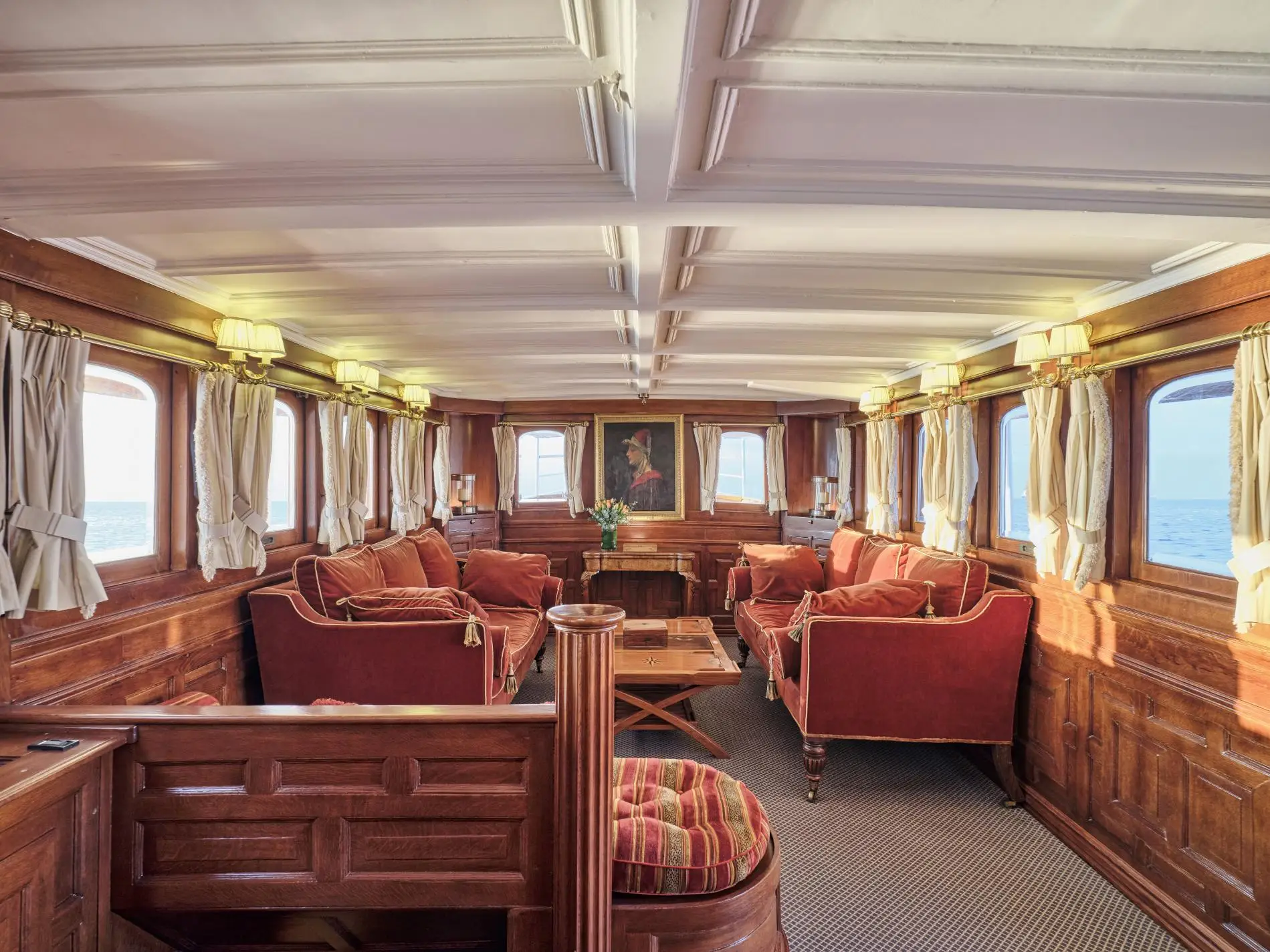 Panelled main saloon with red velvet sofas, arched windows, brass wall lights and a portrait above a fireplace