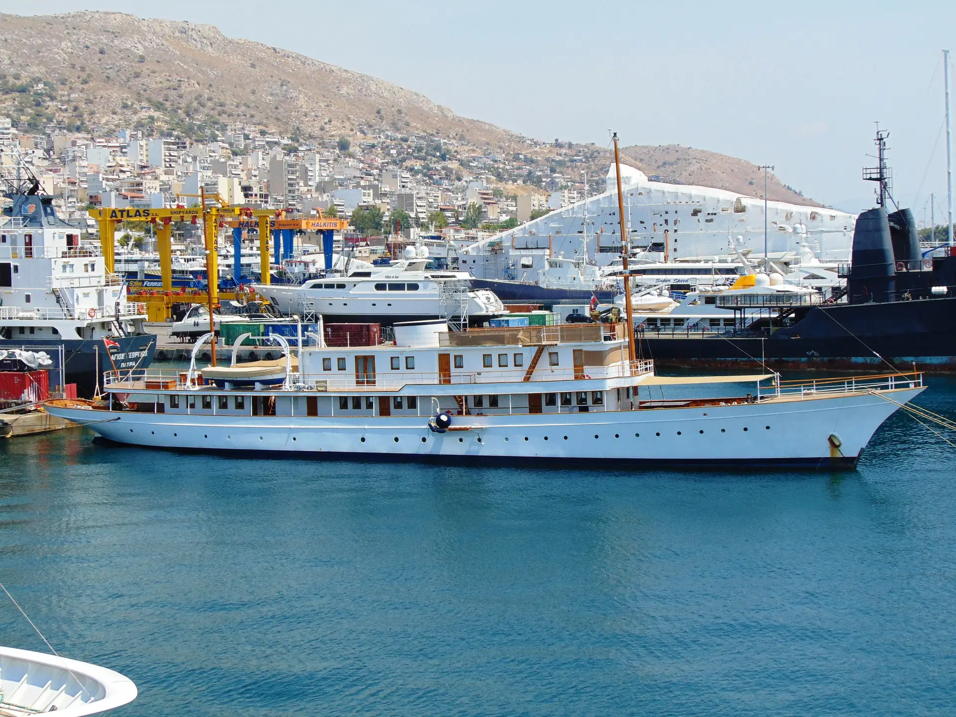Madiz moored alongside in a Greek commercial harbour, with shipyard cranes and a hillside town behind