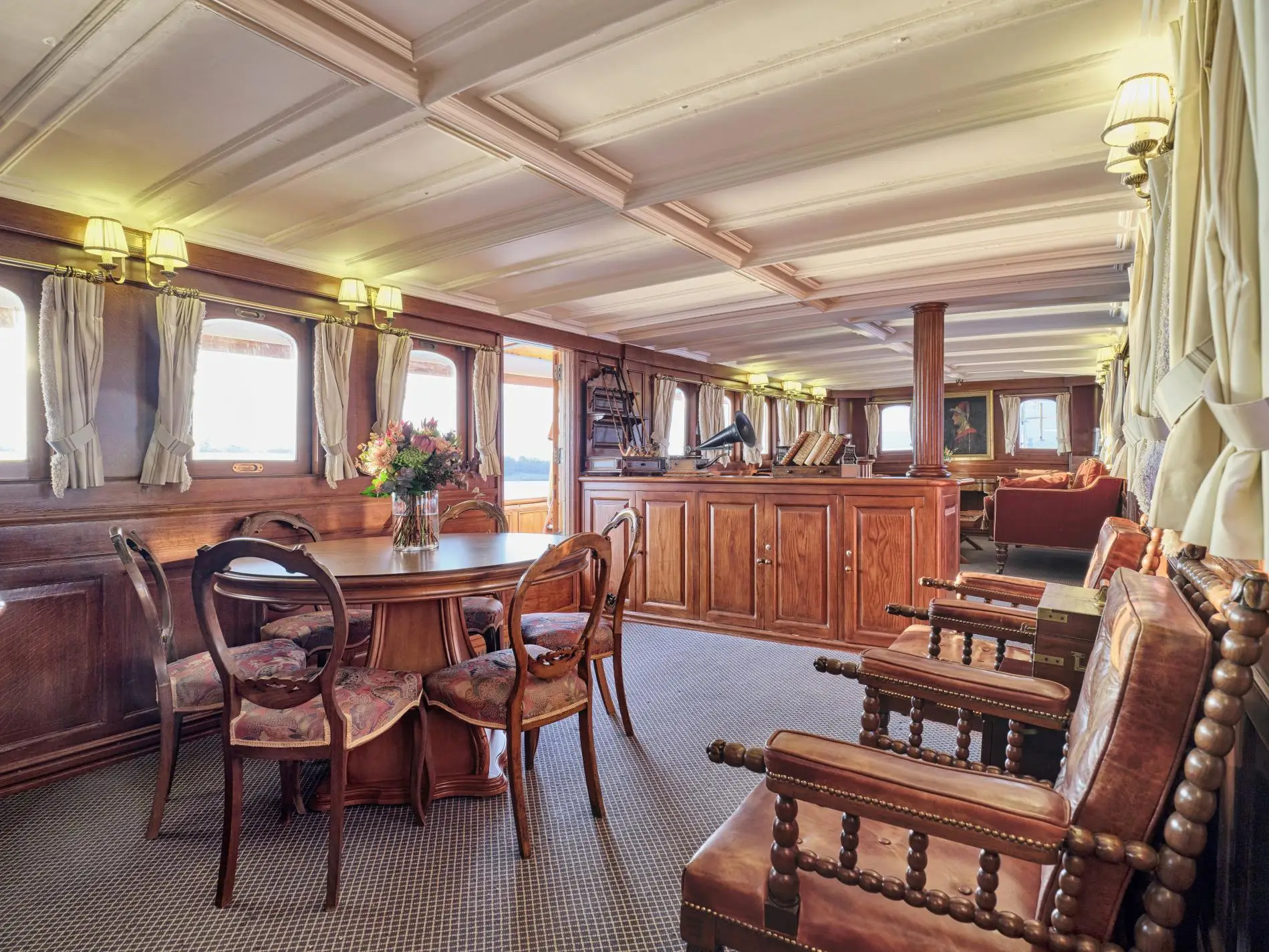 Saloon interior with a round dining table, balloon-back chairs, leather armchairs and a horn gramophone on a sideboard