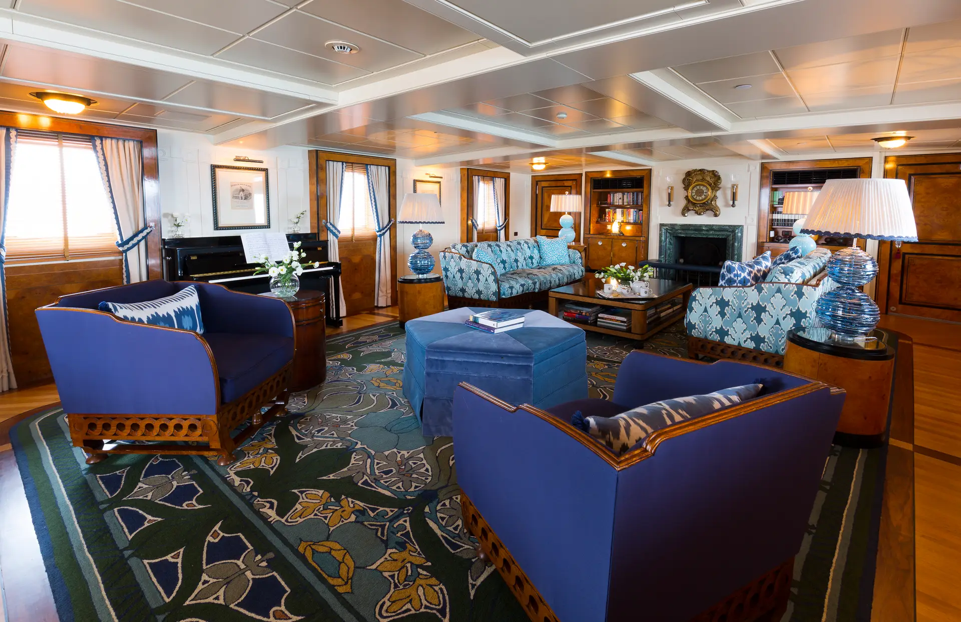 Main saloon with blue upholstered sofas, patterned carpet, upright piano and fireplace