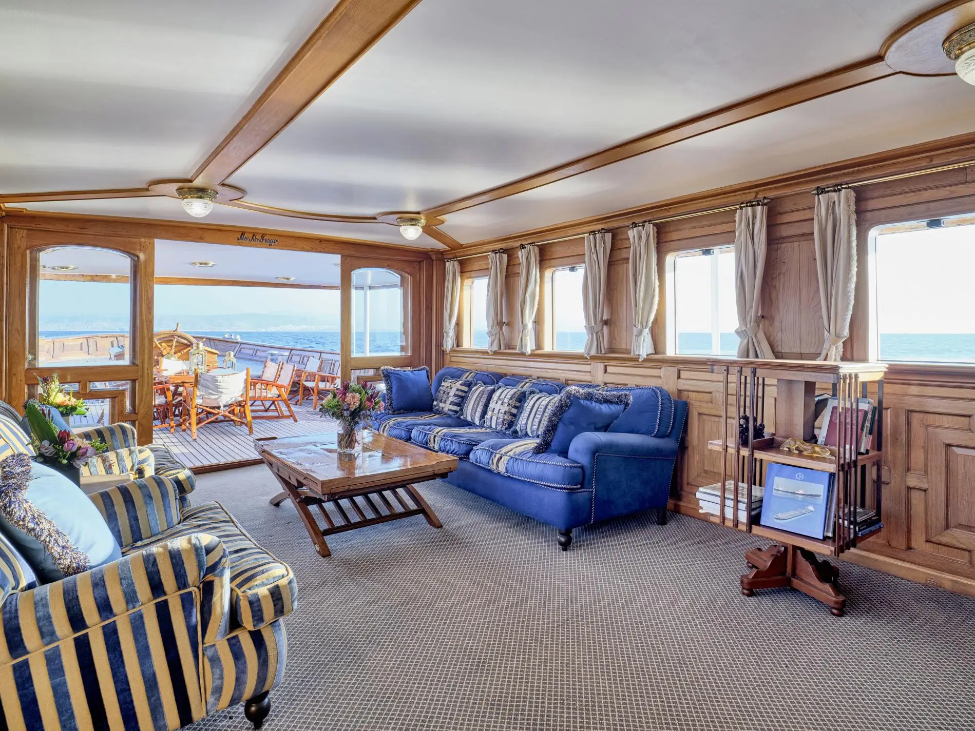 Deckhouse saloon with oak panelling, blue upholstered sofas and open doors onto the teak deck