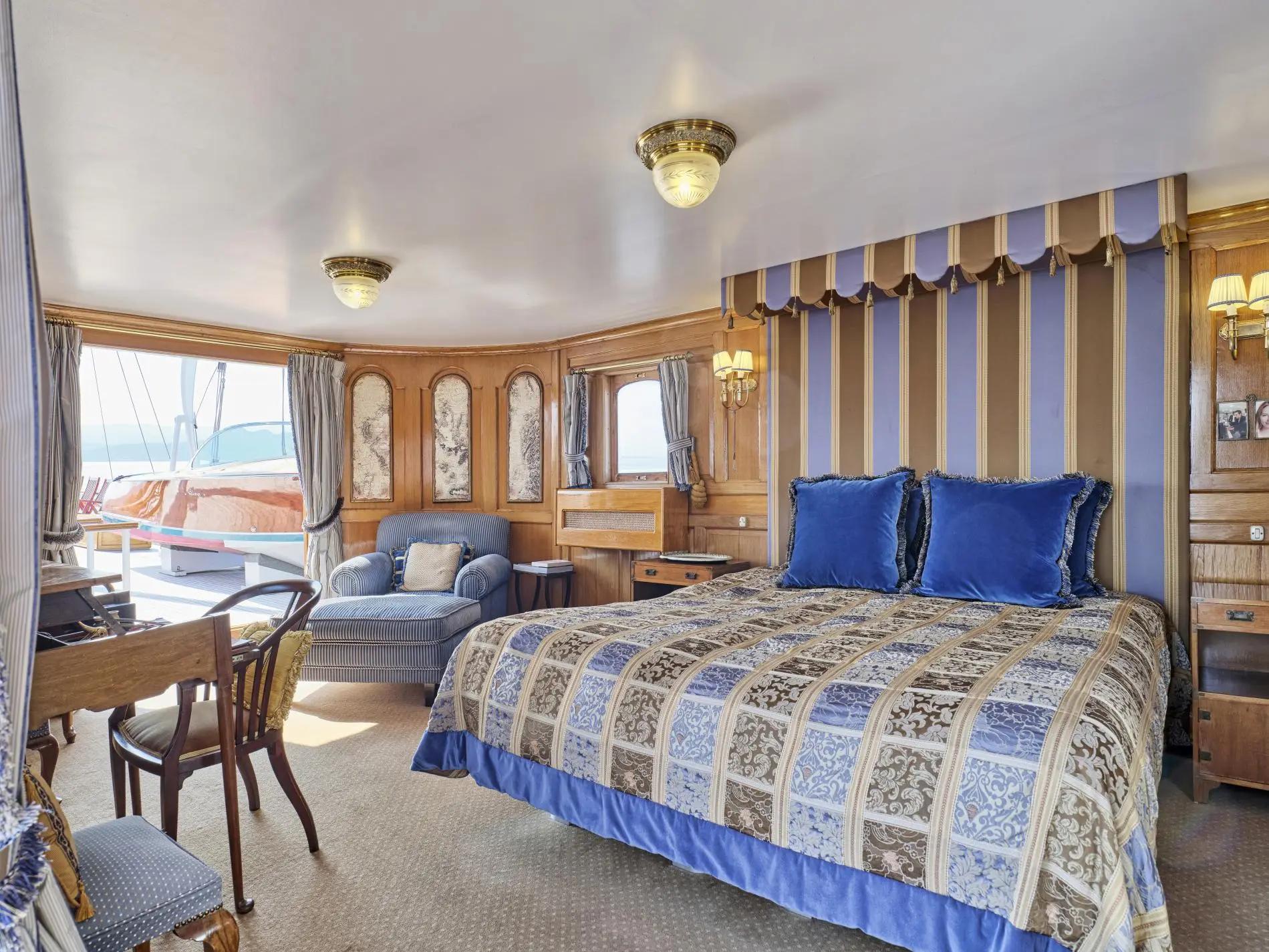 Guest cabin with a canopied bed in blue and gold fabrics, a striped armchair and a window onto the deck