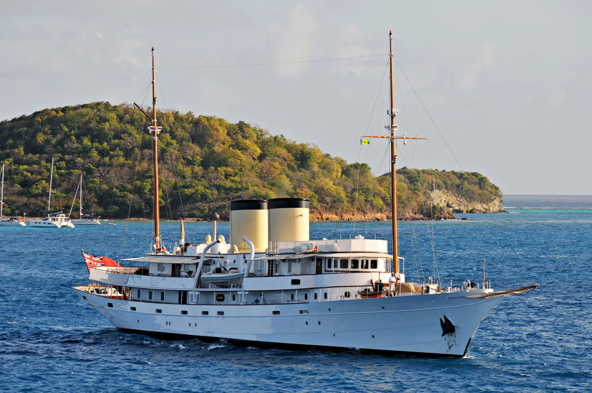 Talitha under way off a wooded headland with a red ensign flying
