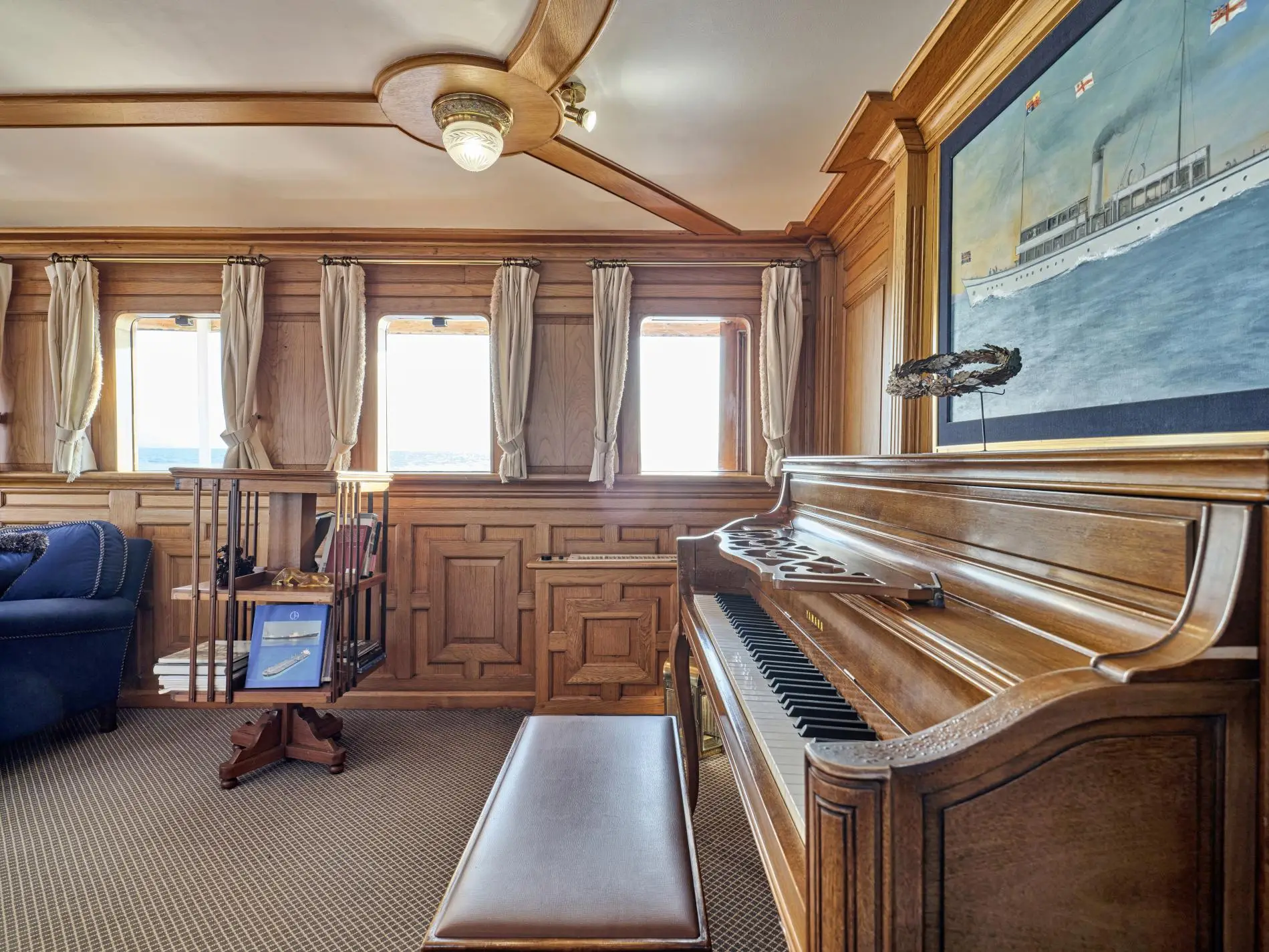 Corner of a panelled saloon with an upright piano and a framed painting of a steam yacht above it